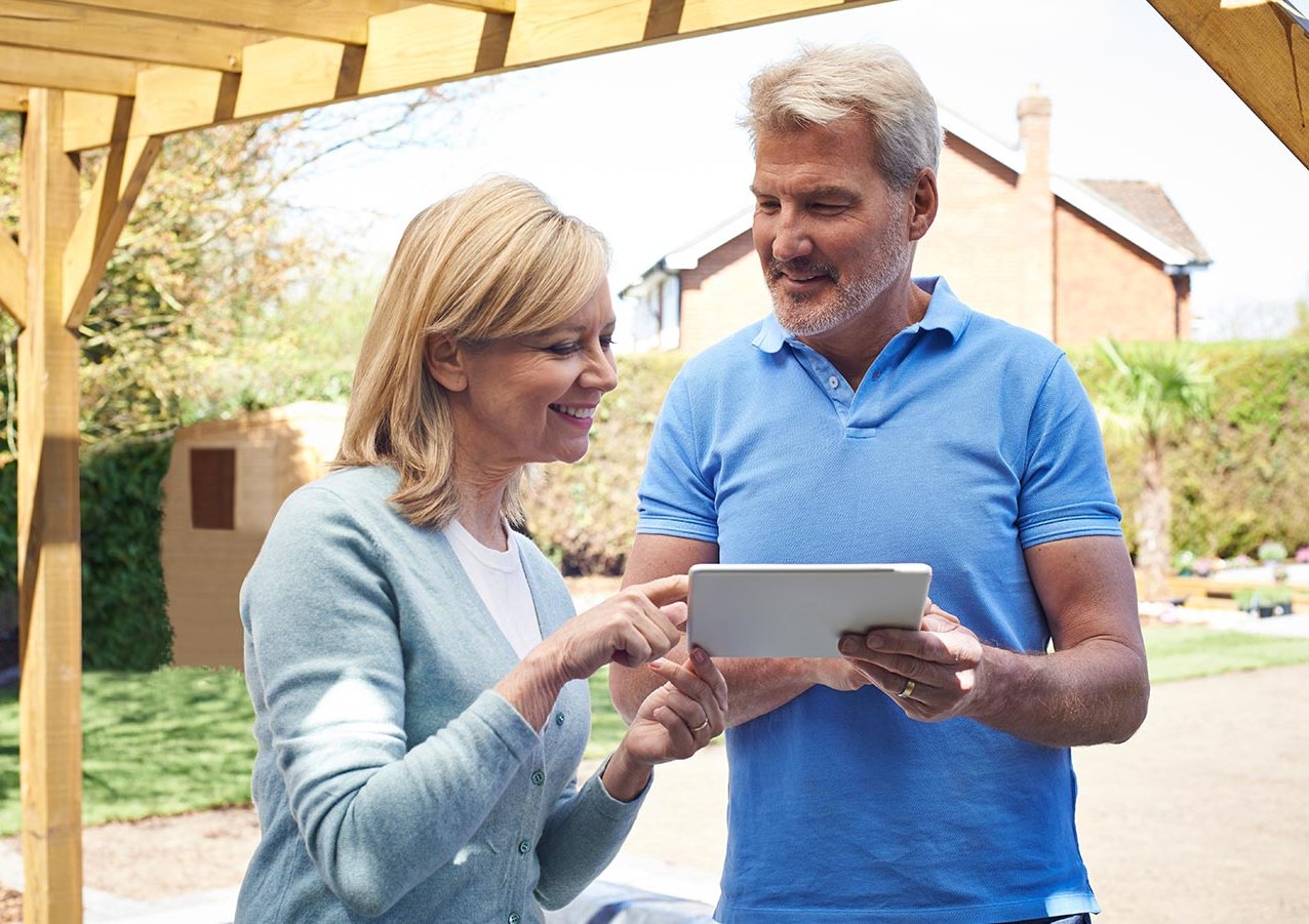 Hand in Hand - Beratung, Lieferung und Montage von Gartenhäusern, Terrassen und Zaun Ein Mann und eine Frau stehen unter einer Terrassenüberdachung mit einem Tablet in der Hand